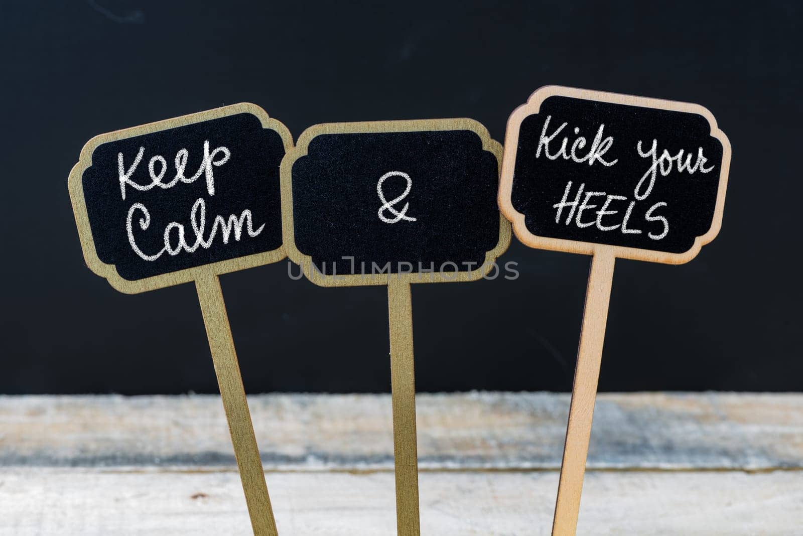 Keep Calm and Kick Your Heels message written with chalk on mini blackboard labels, defocused chalkboard and wooden table in background. Fun and humor concept
