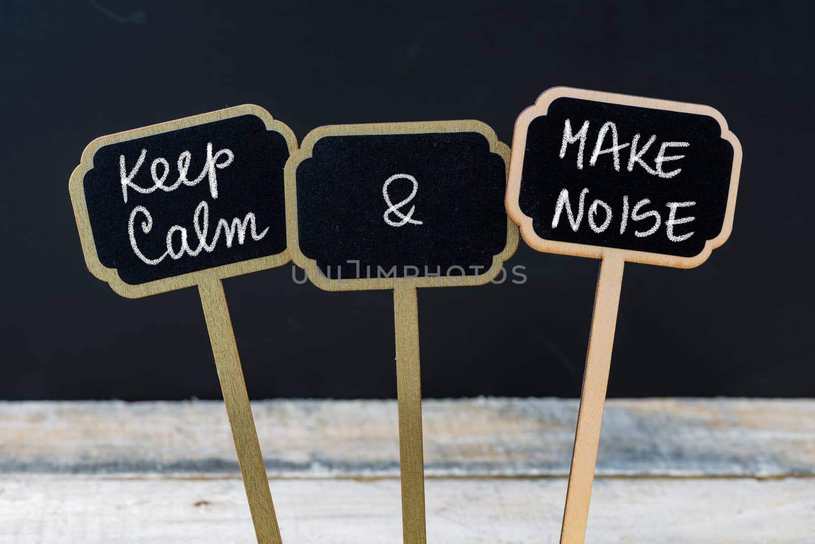 Keep Calm and Make Noise message written with chalk on mini blackboard labels, defocused chalkboard and wooden table in background. Fun and humor concept