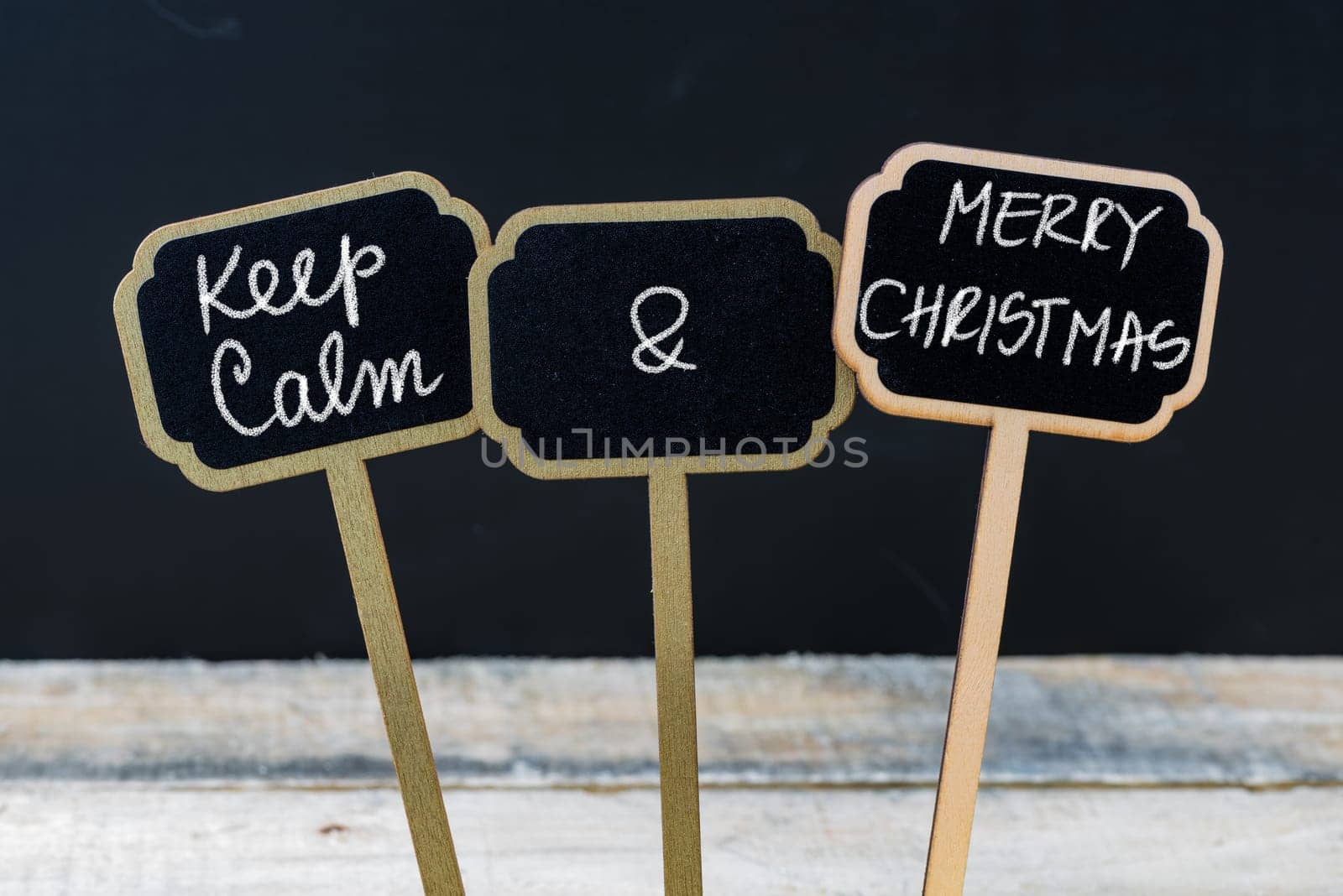 Keep Calm and Merry Christmas message written with chalk on mini blackboard labels, defocused chalkboard and wooden table in background. Fun and humor concept