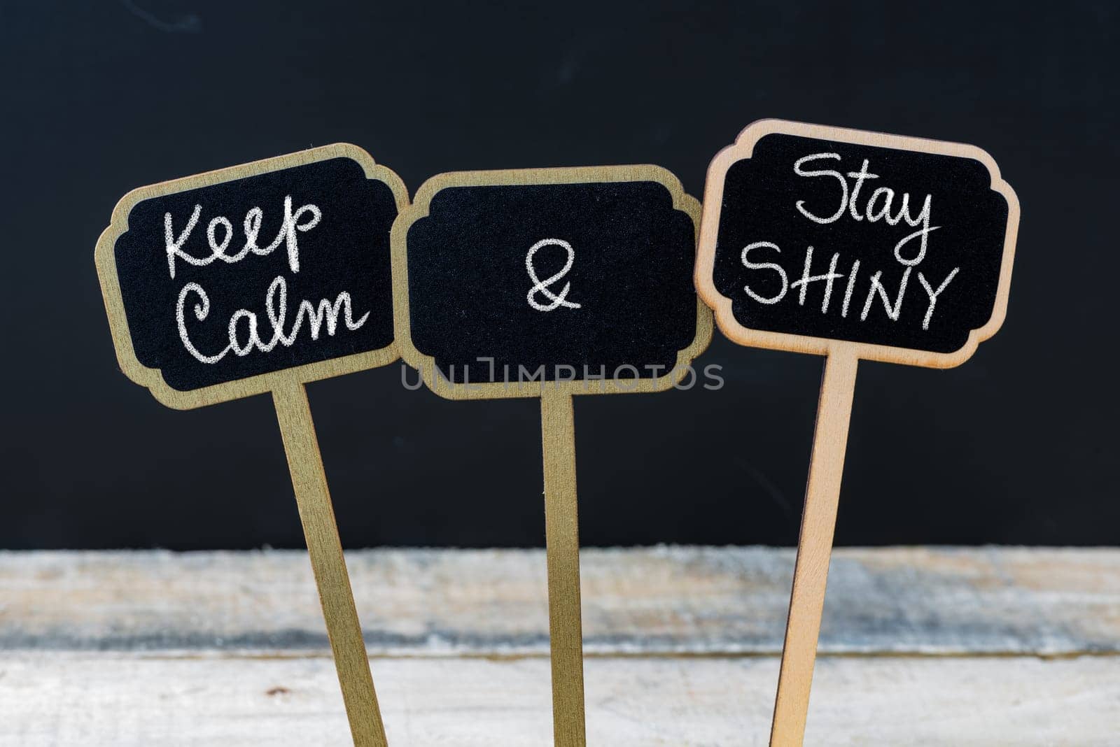 Keep Calm and Stay Shiny message written with chalk on mini blackboard labels, defocused chalkboard and wooden table in background. Fun and humor concept