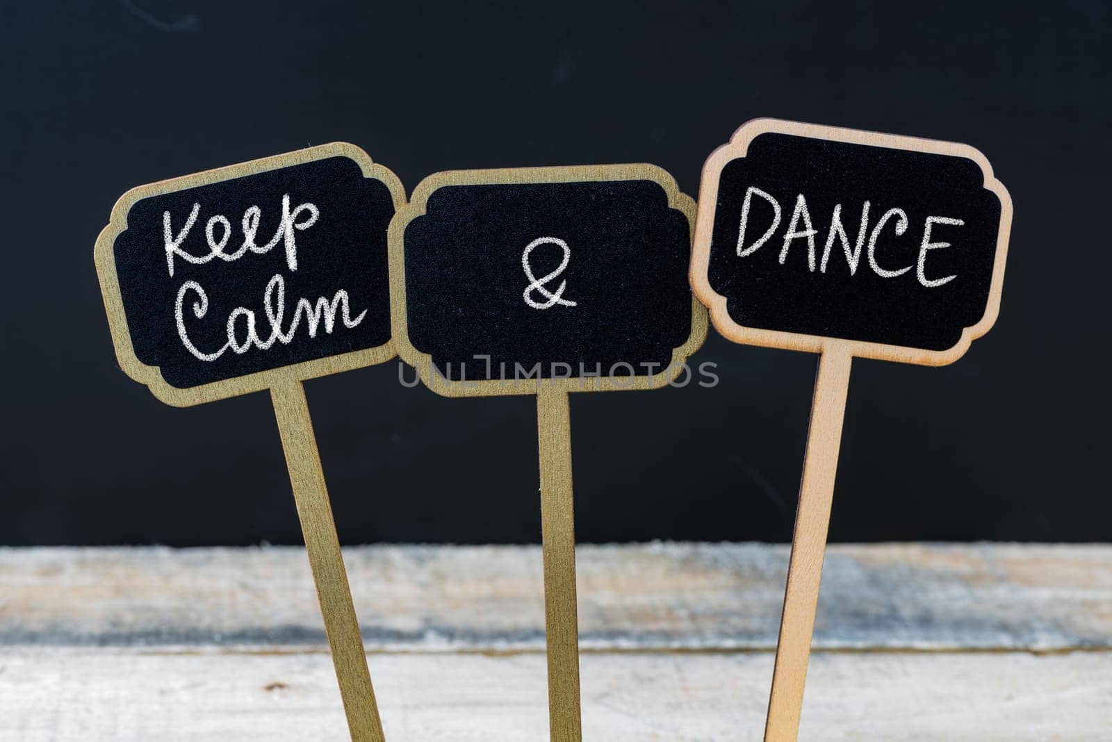 Keep Calm and Dance message written with chalk on mini blackboard labels, defocused chalkboard and wooden table in background. Fun and humor concept