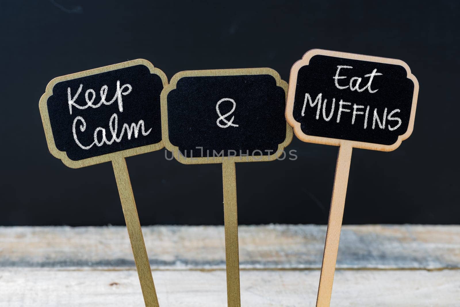 Keep Calm and Eat Muffins message written with chalk on mini blackboard labels, defocused chalkboard and wooden table in background. Fun and humor concept