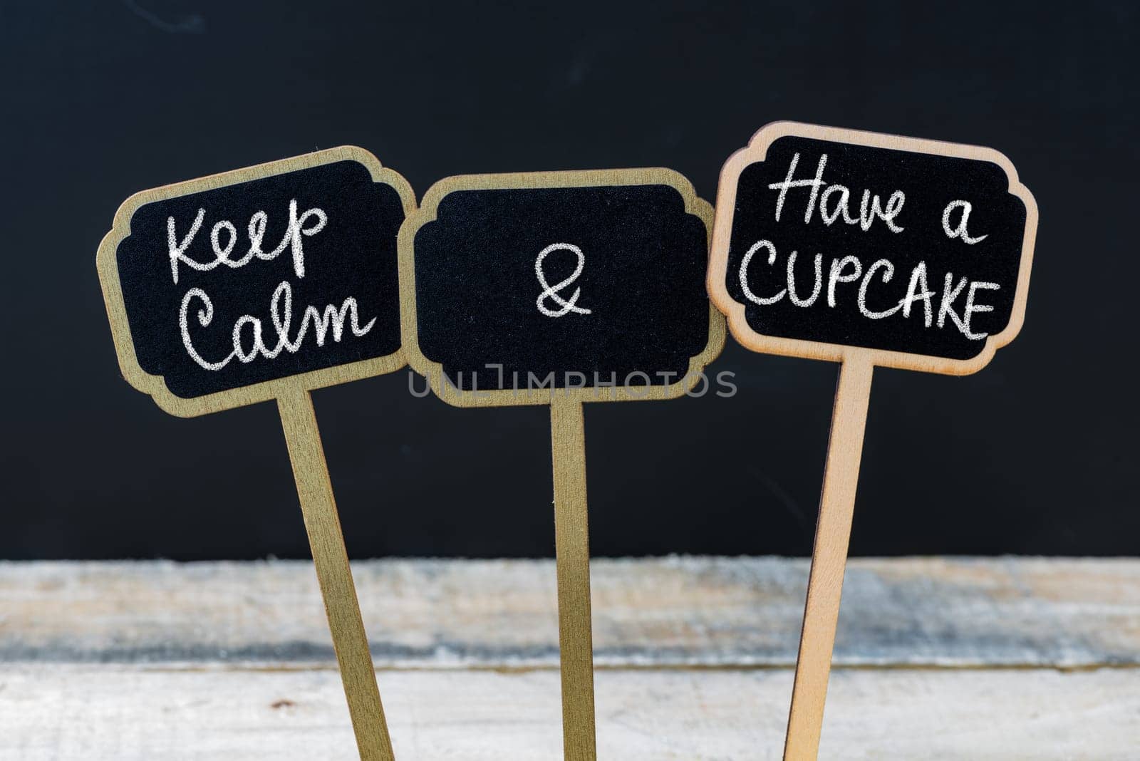 Keep Calm and Have A Cupcake message written with chalk on mini blackboard labels, defocused chalkboard and wooden table in background. Fun and humor concept