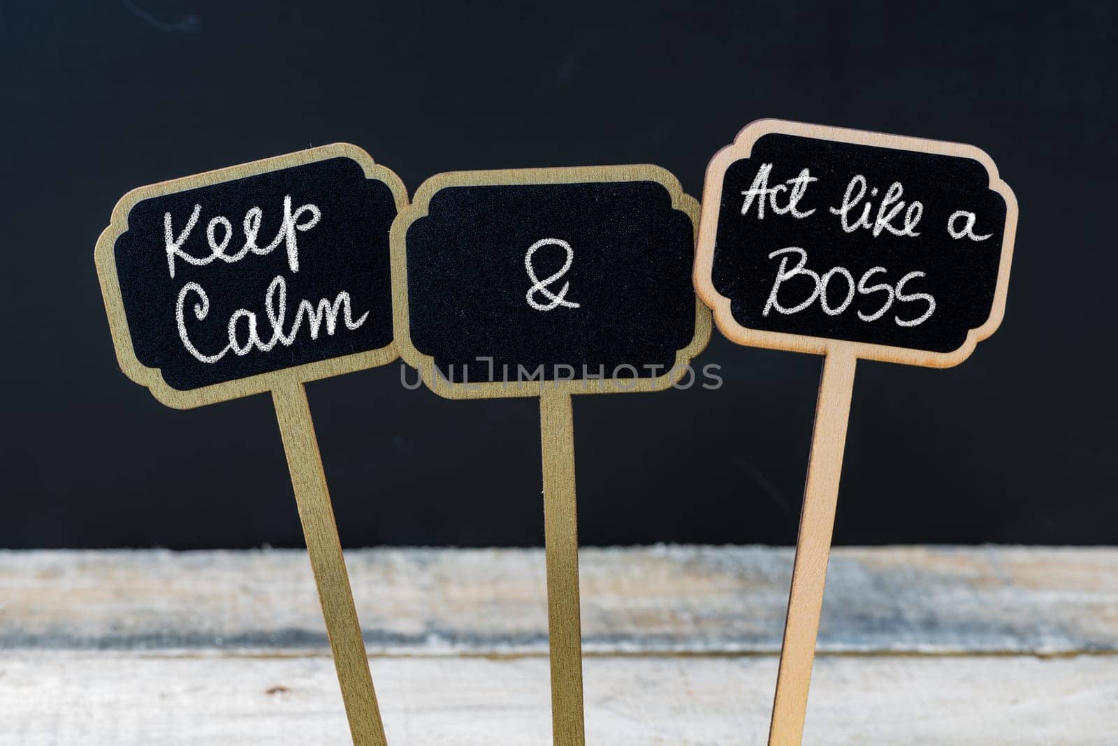 Keep Calm and Act Like A Boss message written with chalk on mini blackboard labels, defocused chalkboard and wooden table in background. Fun and humor concept