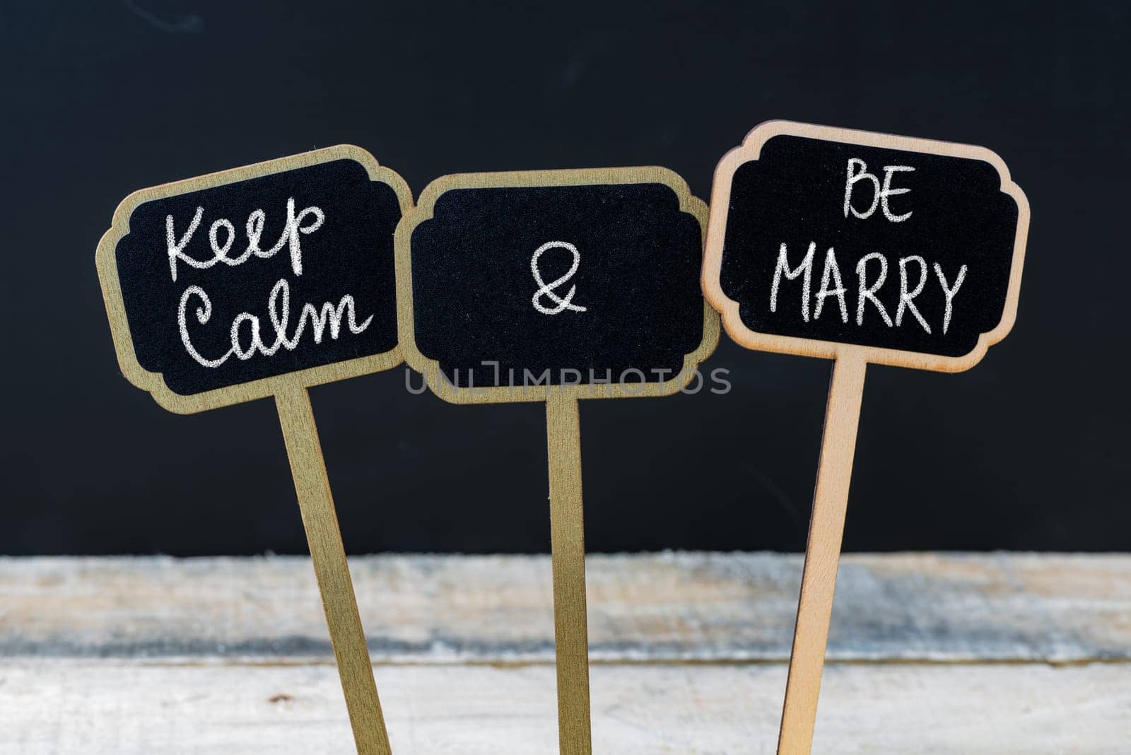 Keep Calm and Be Marry message written with chalk on mini blackboard labels, defocused chalkboard and wooden table in background. Fun and humor concept