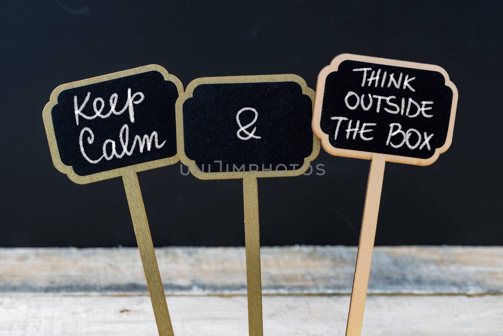 Keep Calm and Think Outside The Box message written with chalk on mini blackboard labels, defocused chalkboard and wooden table in background. Fun and humor concept