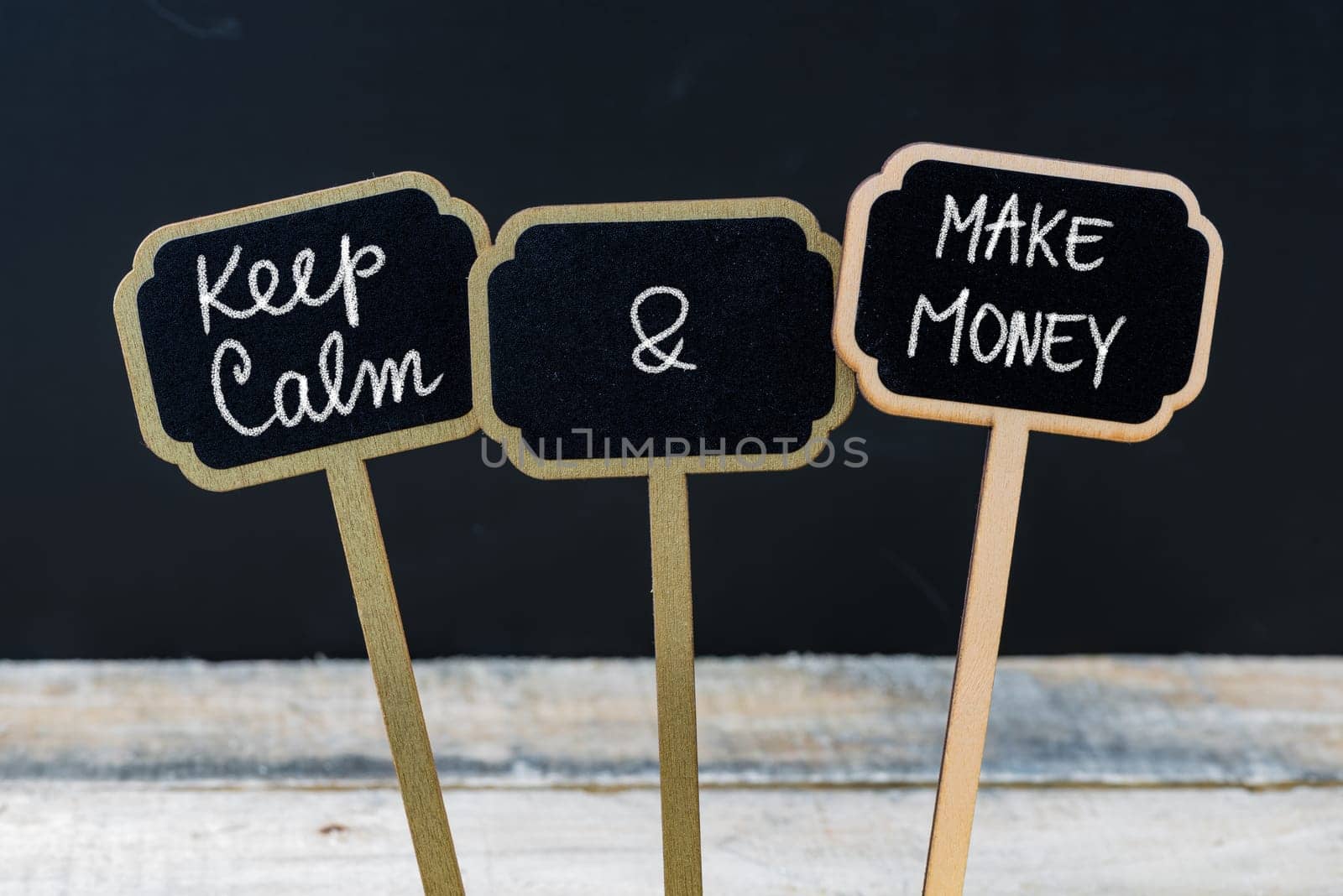 Keep Calm and Make Money message written with chalk on mini blackboard labels, defocused chalkboard and wooden table in background. Fun and humor concept