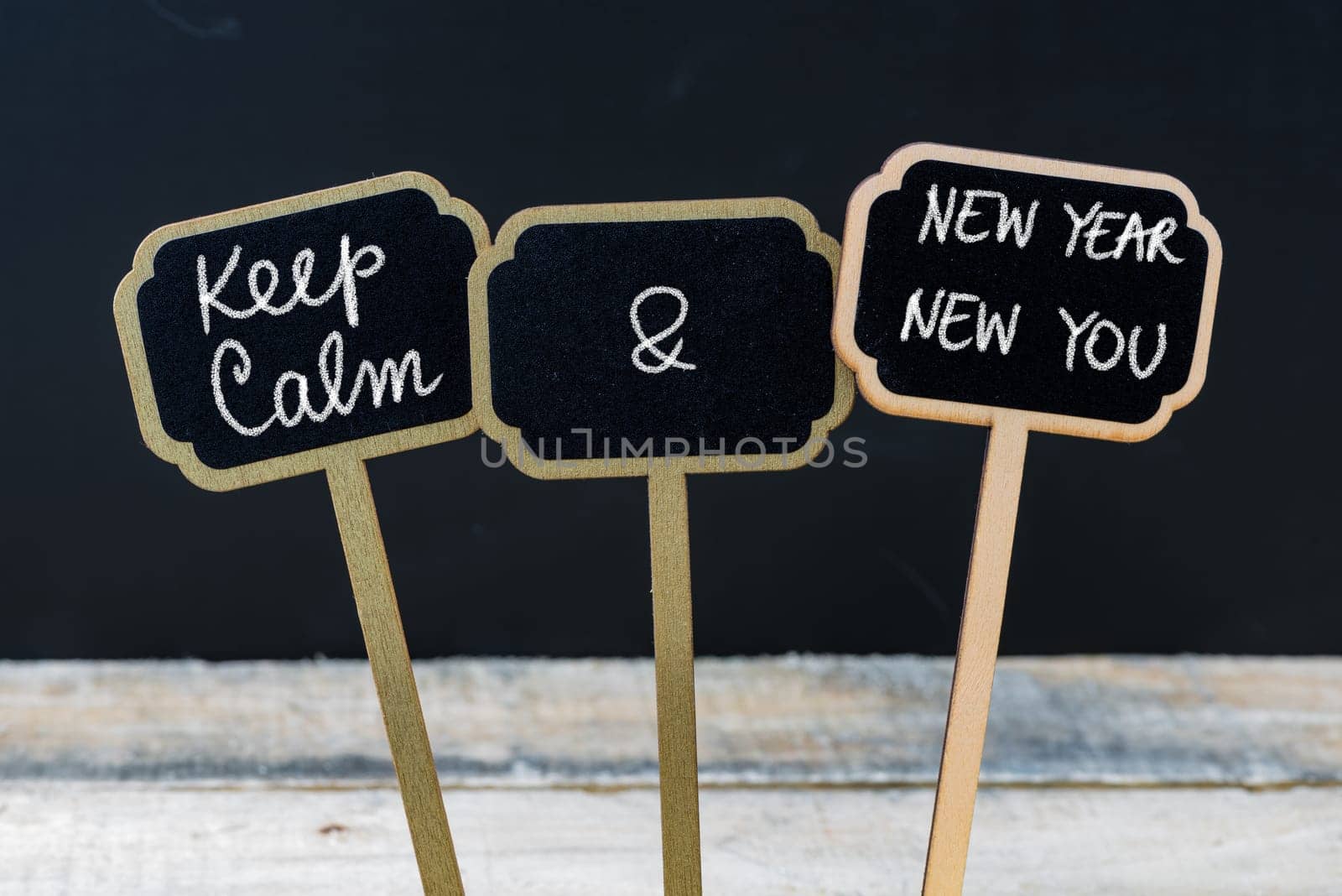 Keep Calm and New Year New You message written with chalk on mini blackboard labels, defocused chalkboard and wooden table in background. Fun and humor concept