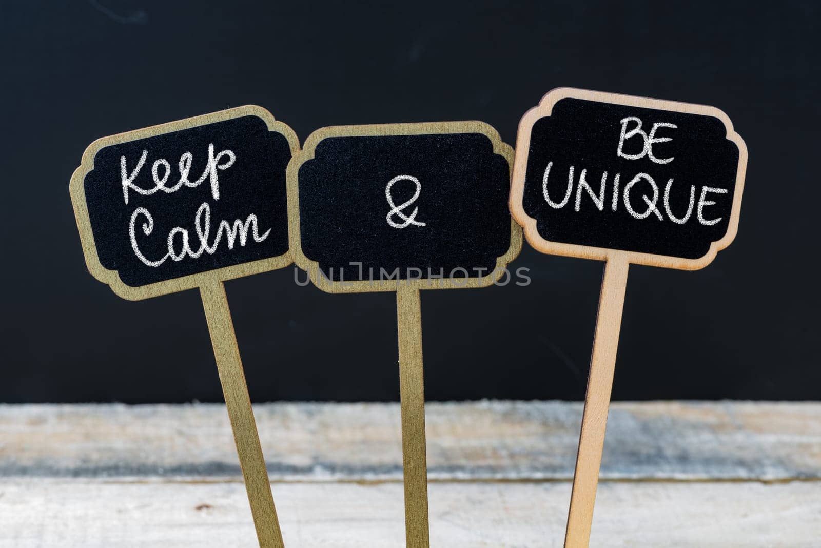 Keep Calm and Be Unique message written with chalk on mini blackboard labels, defocused chalkboard and wooden table in background. Fun and humor concept