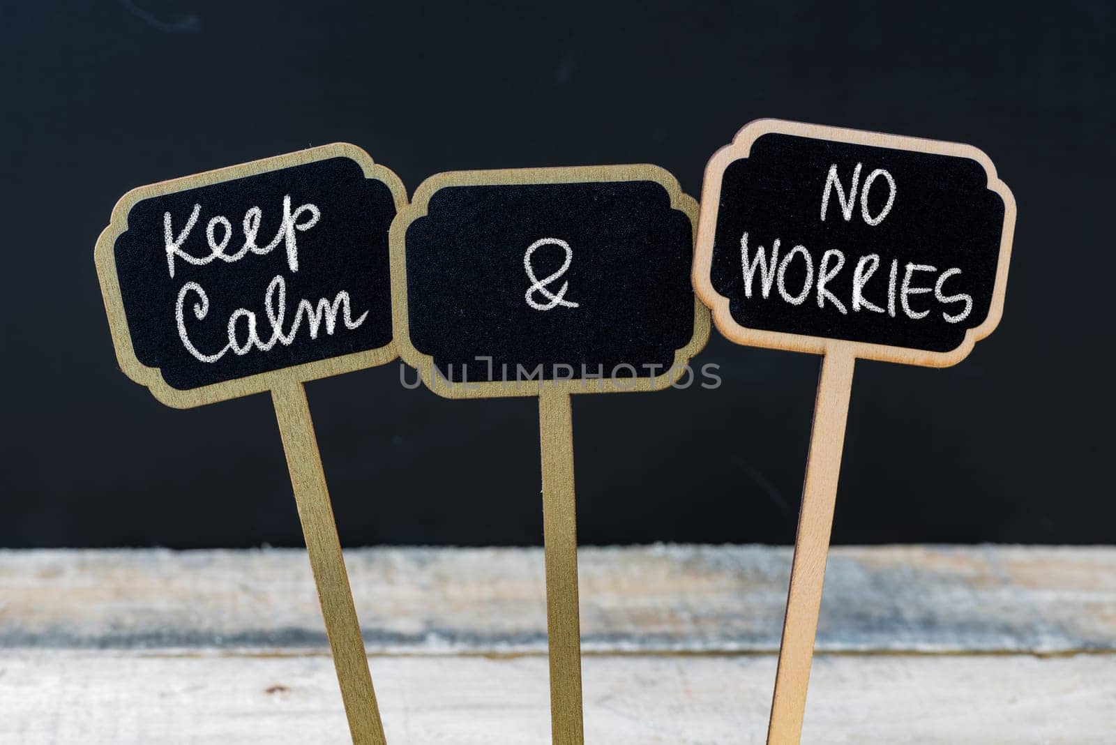 Keep Calm and No Worries message written with chalk on mini blackboard labels, defocused chalkboard and wooden table in background. Fun and humor concept