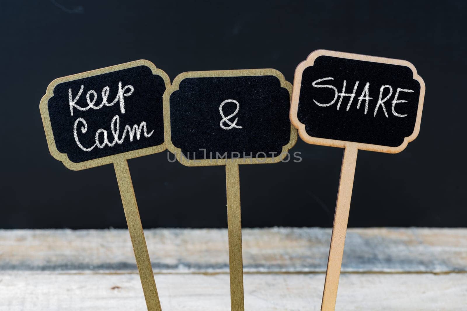 Keep Calm and Share message written with chalk on mini blackboard labels, defocused chalkboard and wooden table in background. Fun and humor concept