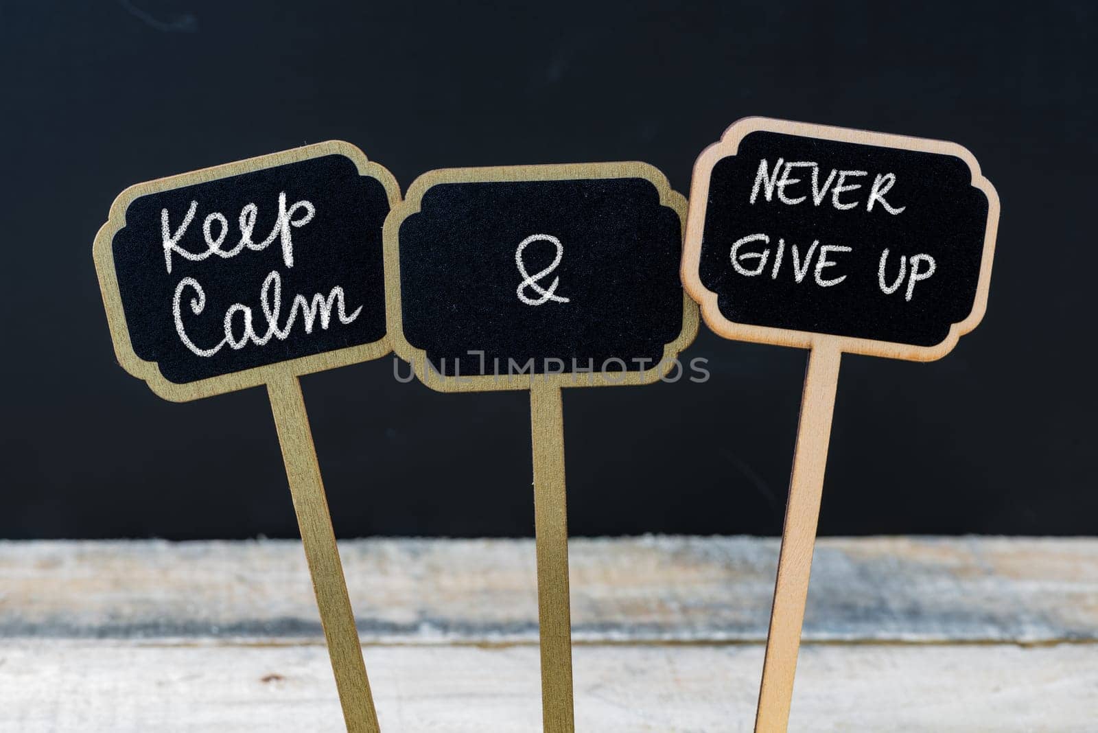 Keep Calm and Never Give Up message written with chalk on mini blackboard labels, defocused chalkboard and wooden table in background. Fun and humor concept