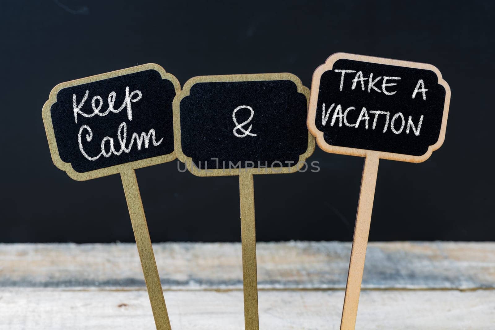 Keep Calm and Take A Vacation message written with chalk on mini blackboard labels, defocused chalkboard and wooden table in background. Fun and humor concept