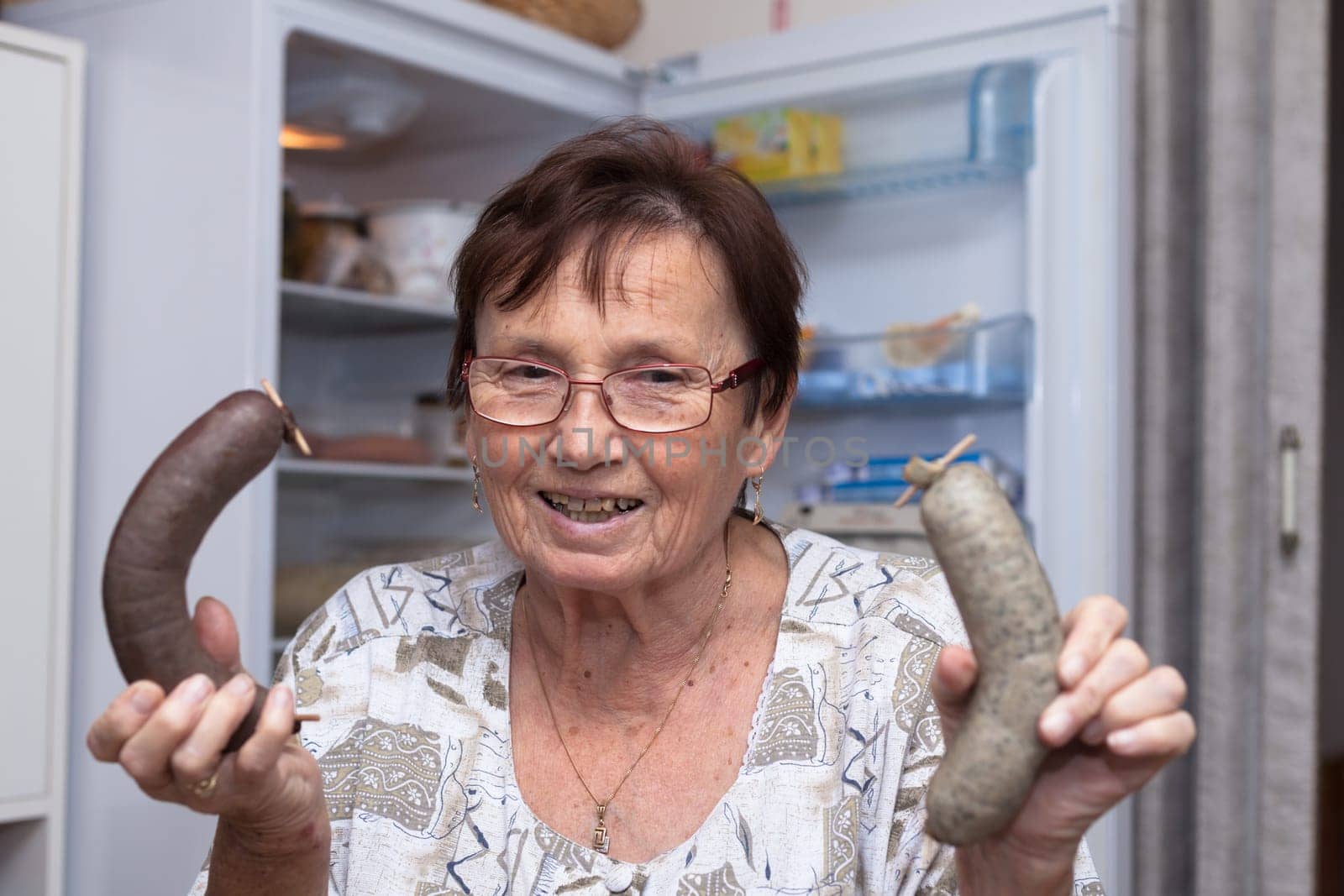 Happy senior woman holding pork liver sausages while standing in front of the open fridge in the kitchen.