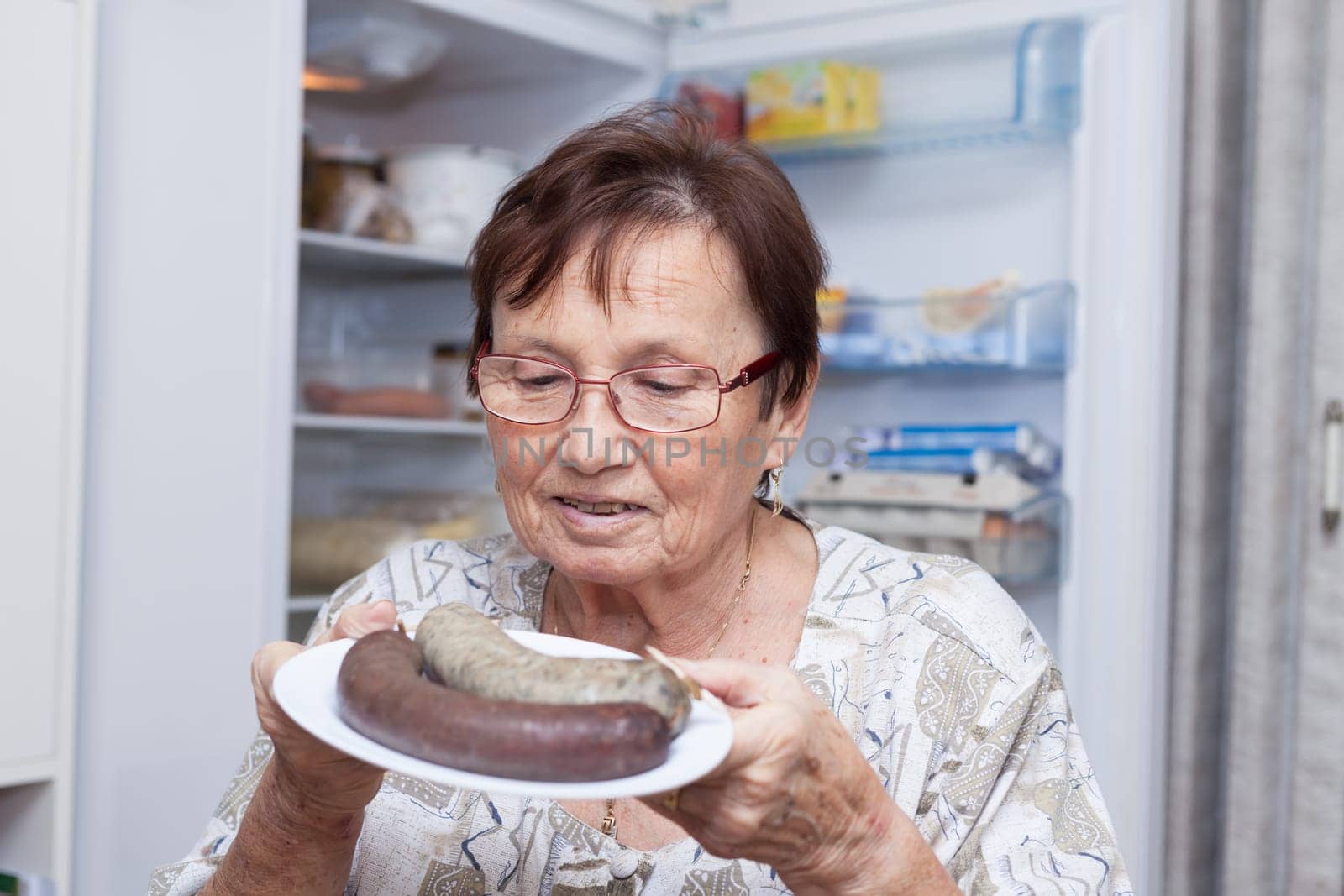 Happy senior woman holding a plate with pork liver sausages while standing in front of the open fridge in the kitchen.