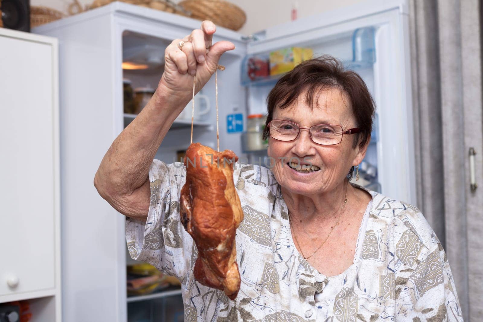Happy senior woman holding smoked meat while standing in front of the open fridge in the kitchen.