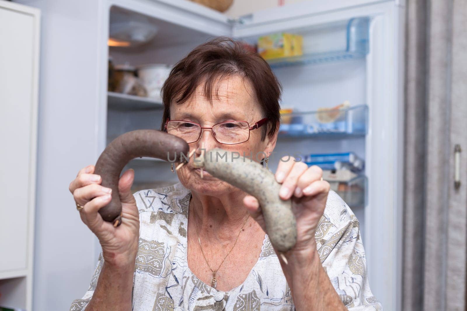 Senior woman holding pork liver sausages while standing in front of the open fridge in the kitchen.