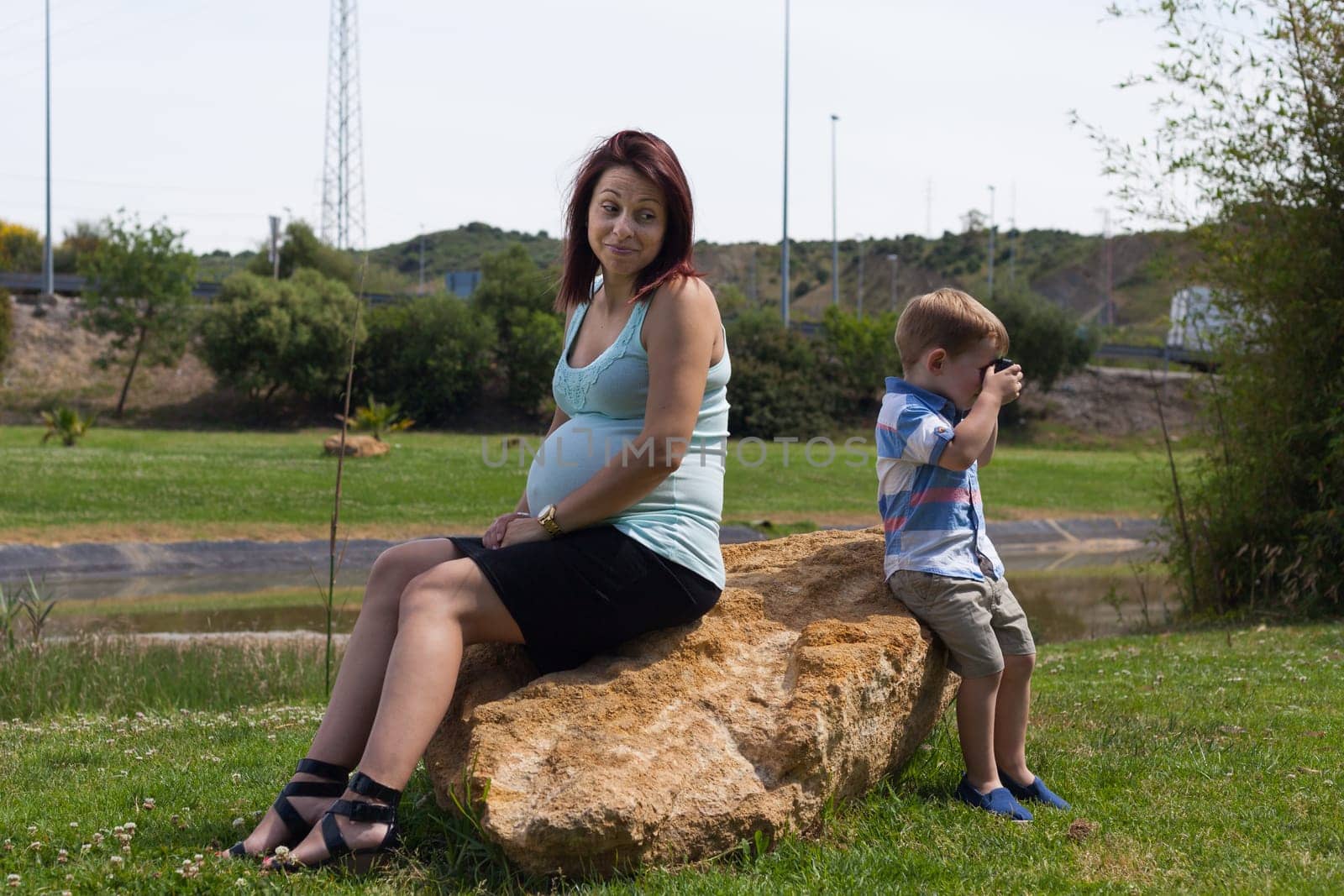 Pregnant woman and child boy sitting back to back outdoors.
