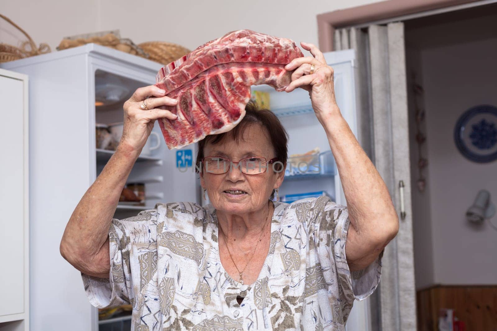 Senior woman holding raw pork ribs while standing in front of the open fridge in the kitchen.