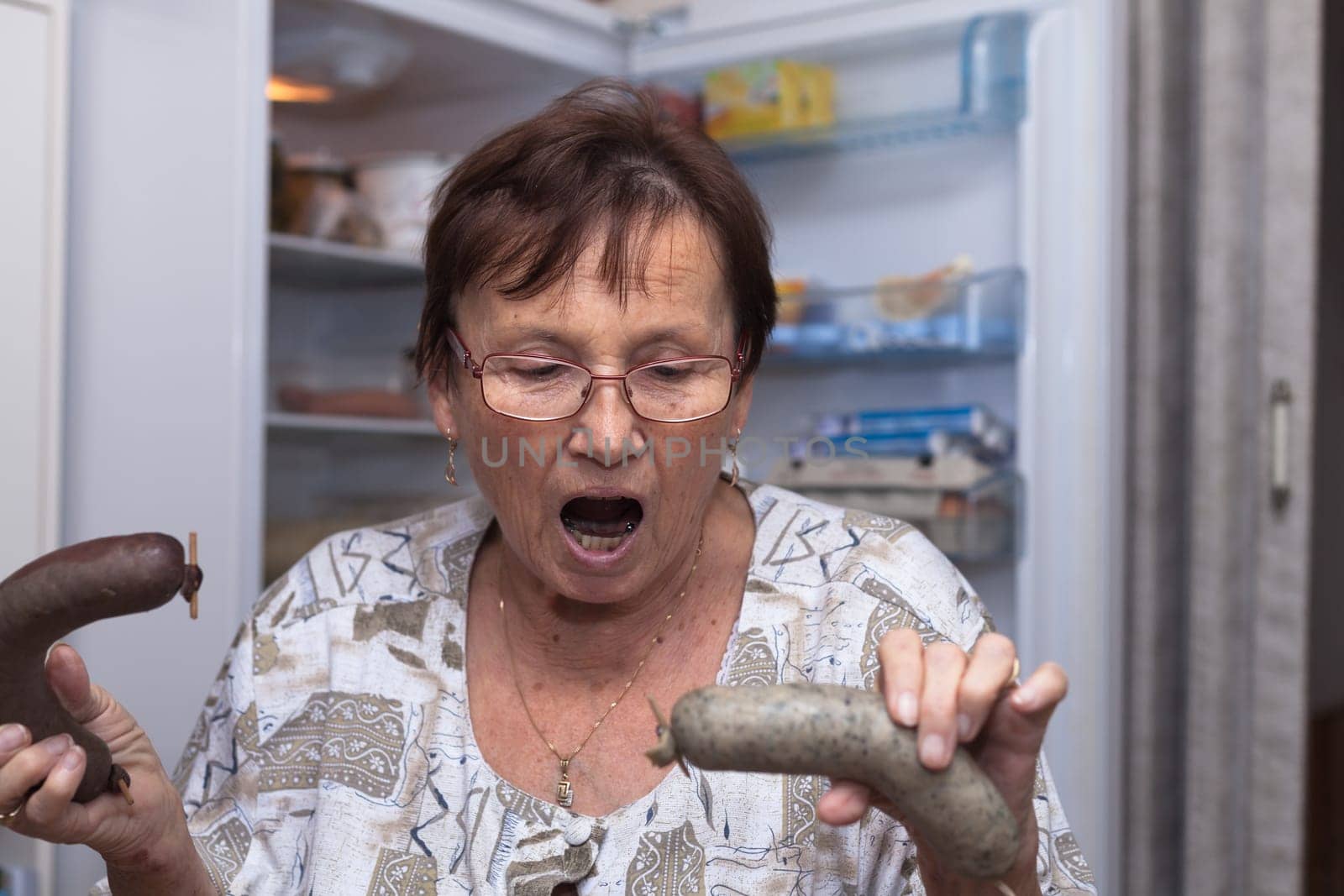 Shocked senior woman holding pork liver sausages while standing in front of the open fridge in the kitchen.