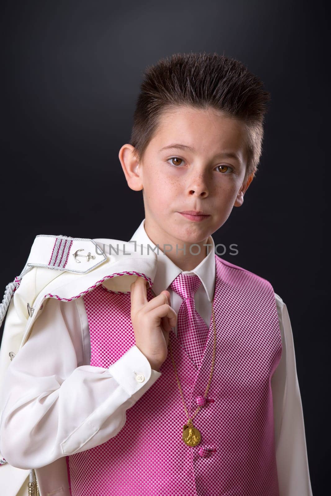 Young boy in his First Holy Communion holding his jacket over his shoulder and looking at camera on a black background.