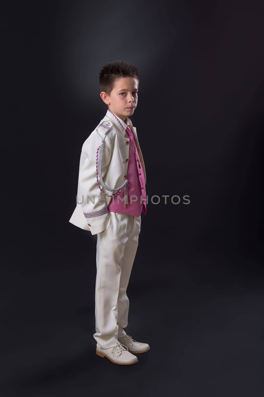 Young boy in his First Holy Communion looking at camera and standing with a serious expression on a black background.
