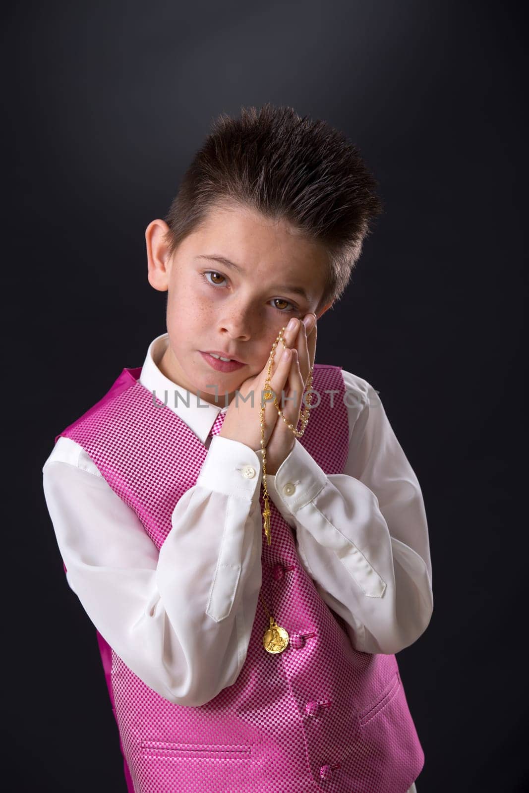 Young boy celebrating his First Holy Communion holding his rosary and looking at camera on a black background.