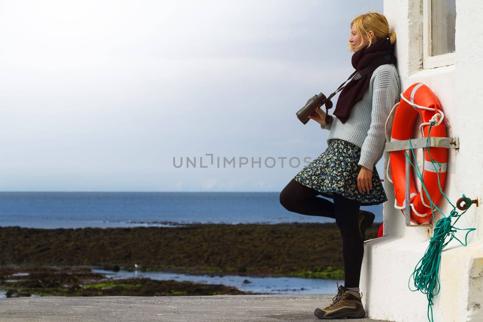 Young Caucasian blonde woman holding binoculars by seaside near a lighthouse with a life buoy