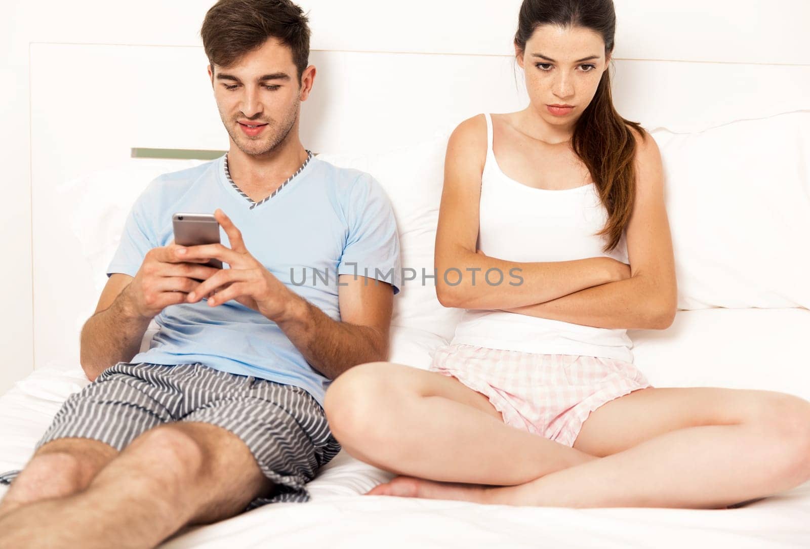 Addicted young man on bed texting while woman looks angry