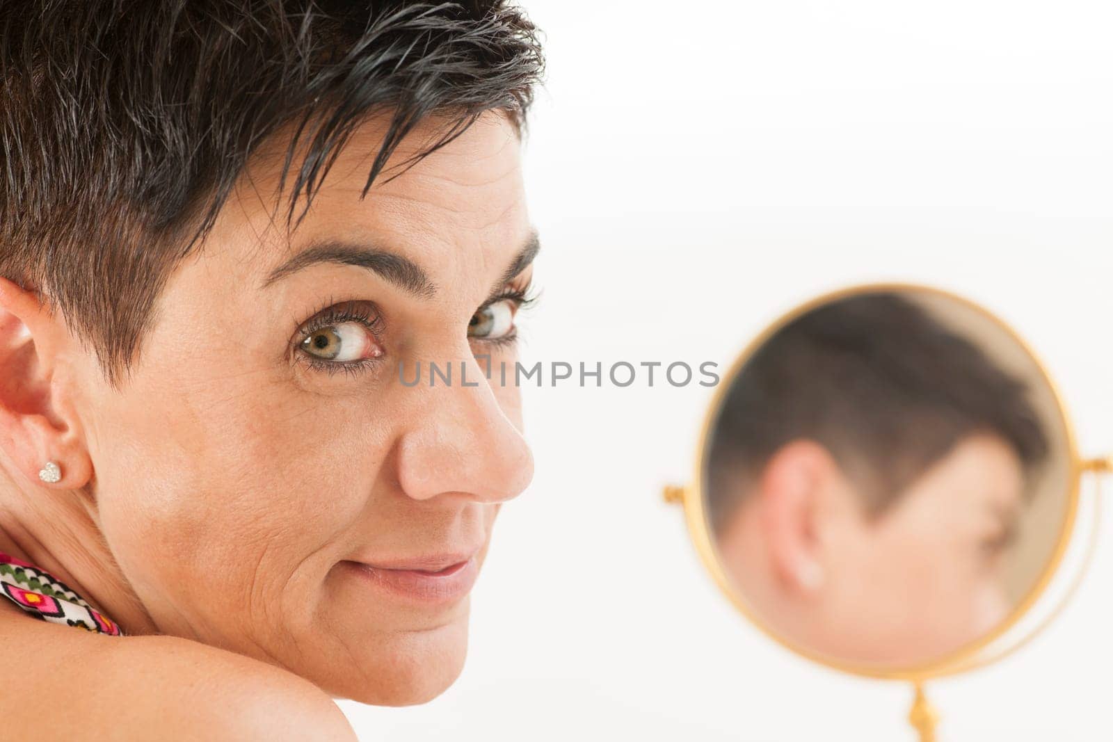 Closeup of a middle-aged black-haired woman with a round make-up mirror against a white background looking into the camera.