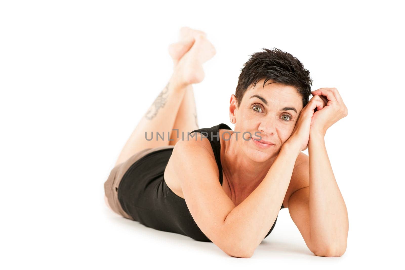 Full-body view of a black-haired middle-aged woman barefoot in shorts and black top lying on her stomach against a white background looking into the camera.
