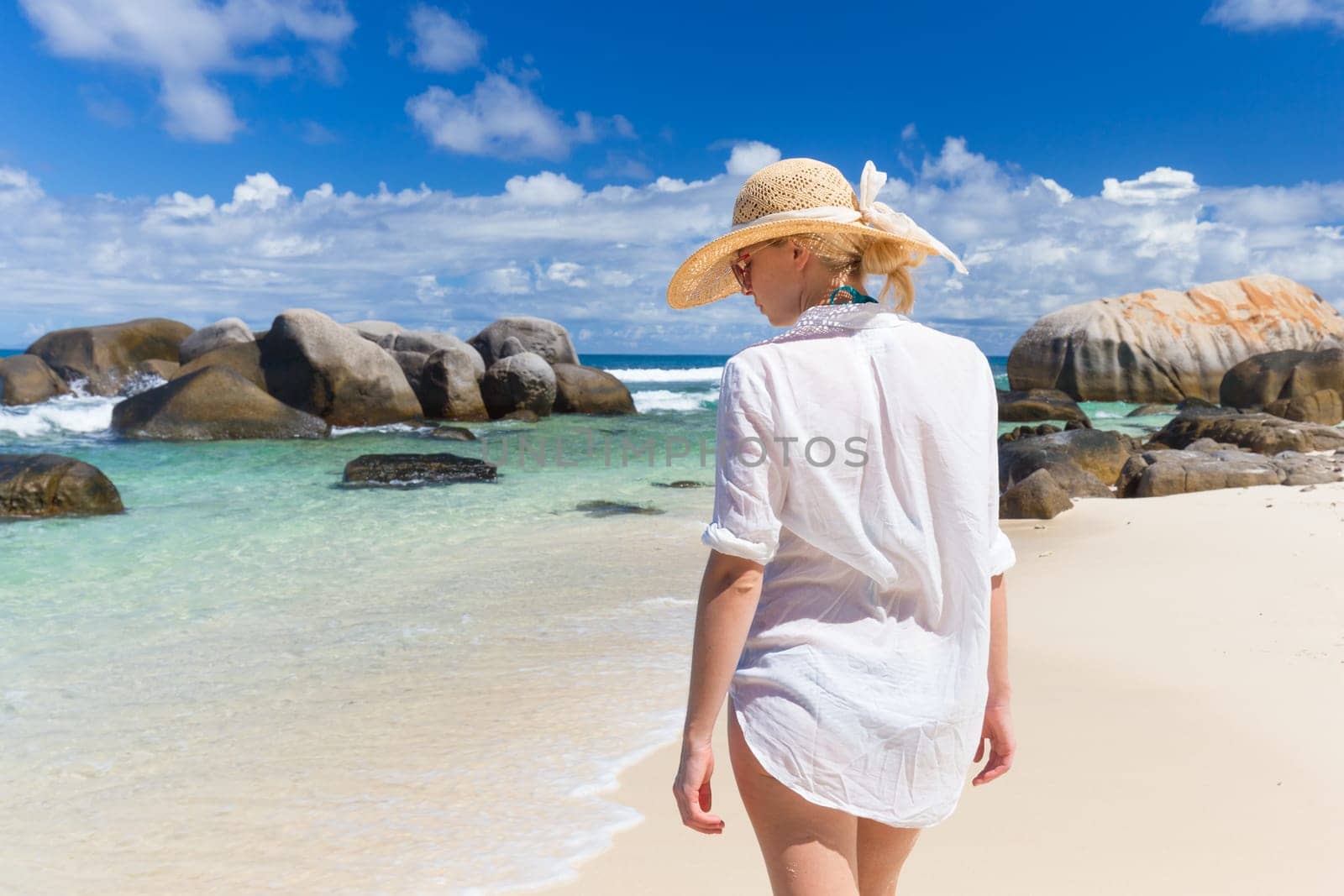 Woman wearing white loose tunic over bikini and beach hat on Mahe Island, Seychelles. Summer vacations on picture perfect tropical beach concept.