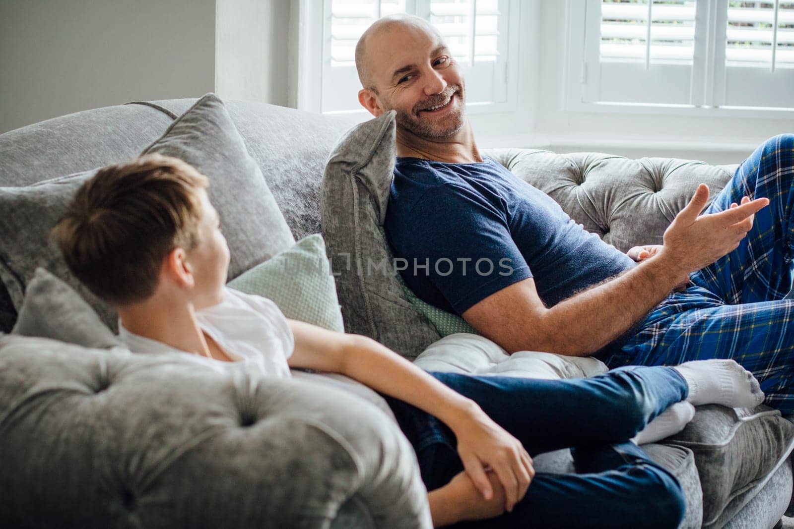 Father and son sitting on the sofa in their home together in teh morning. They are talking and laughing about something they are watching on television.