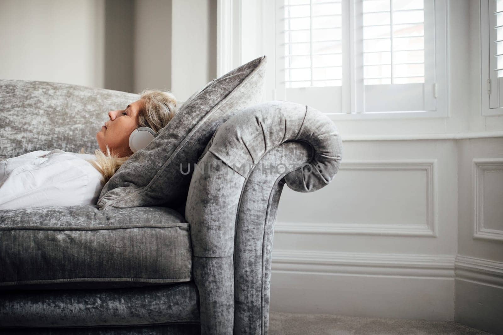 Mature woman lying on her sofa, enjoying listening to some music through headphones.