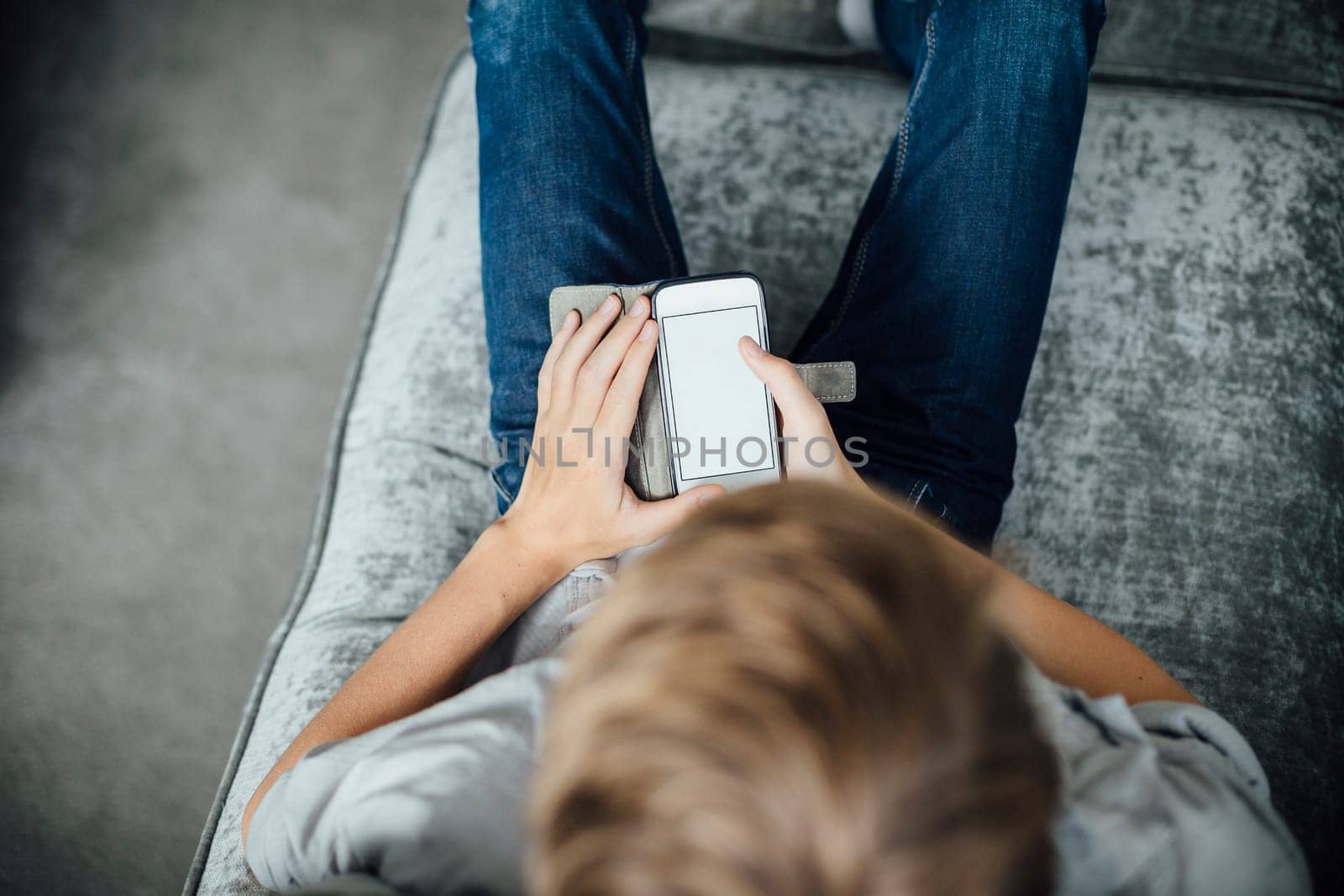 Shot from above of a boy sitting on his sofa at home, using a smartphone.