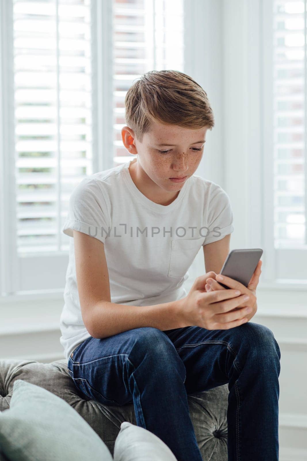 Boy sitting on the sofa in his living room. He is holding a smart phone which he is looking at.