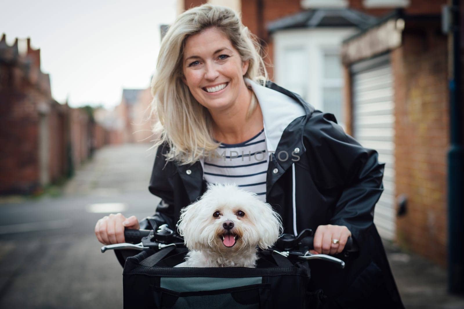 Bichon Frise sitting in a basket on the front of it's owners bike. It is looking at the camera with it's tongue out. The woman is out of focus in the background, smiling at the camera.