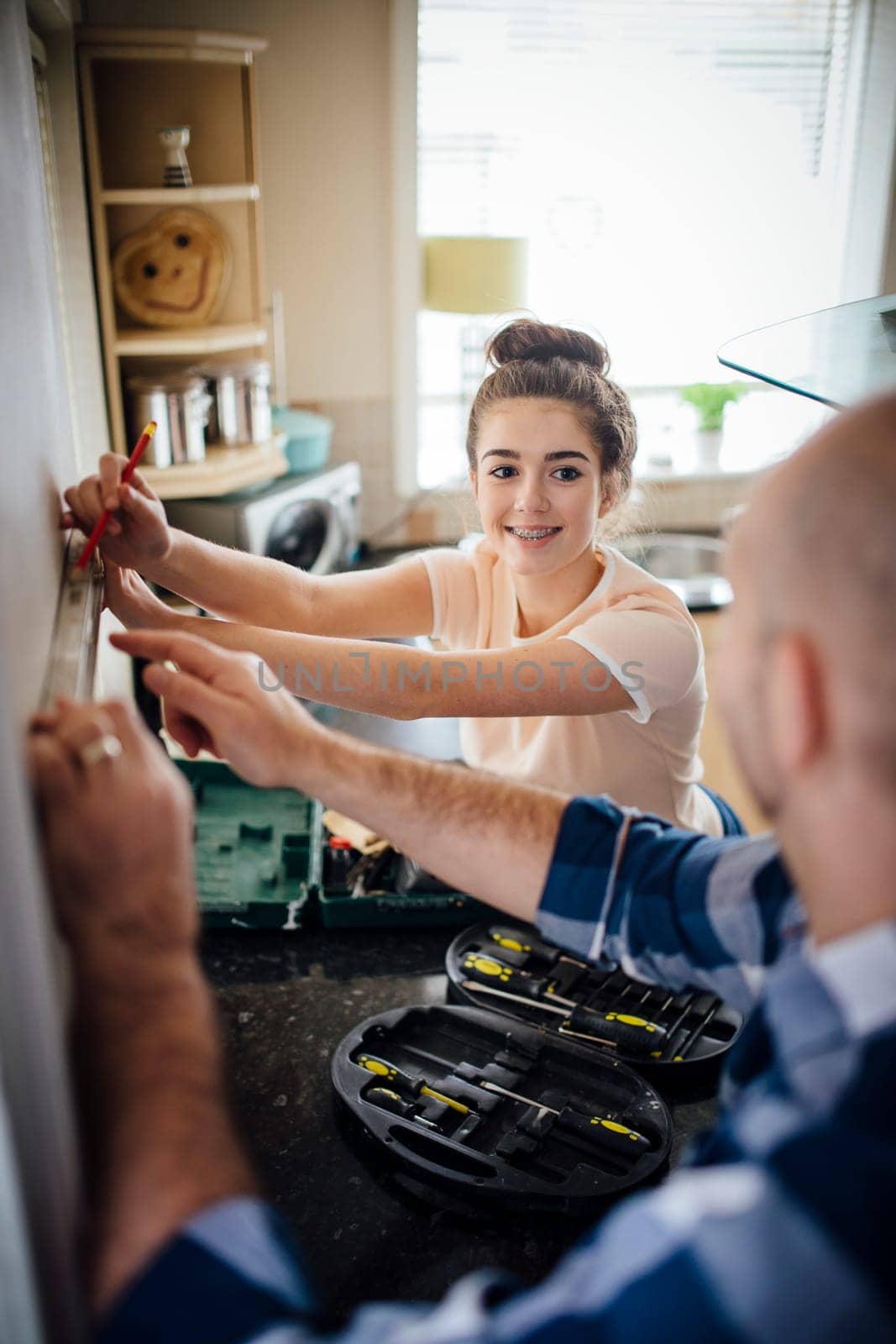 Teenage girl helping her father with DIy in their kitchen. The girl is marking the wall with a pencil, while smiling at her father.