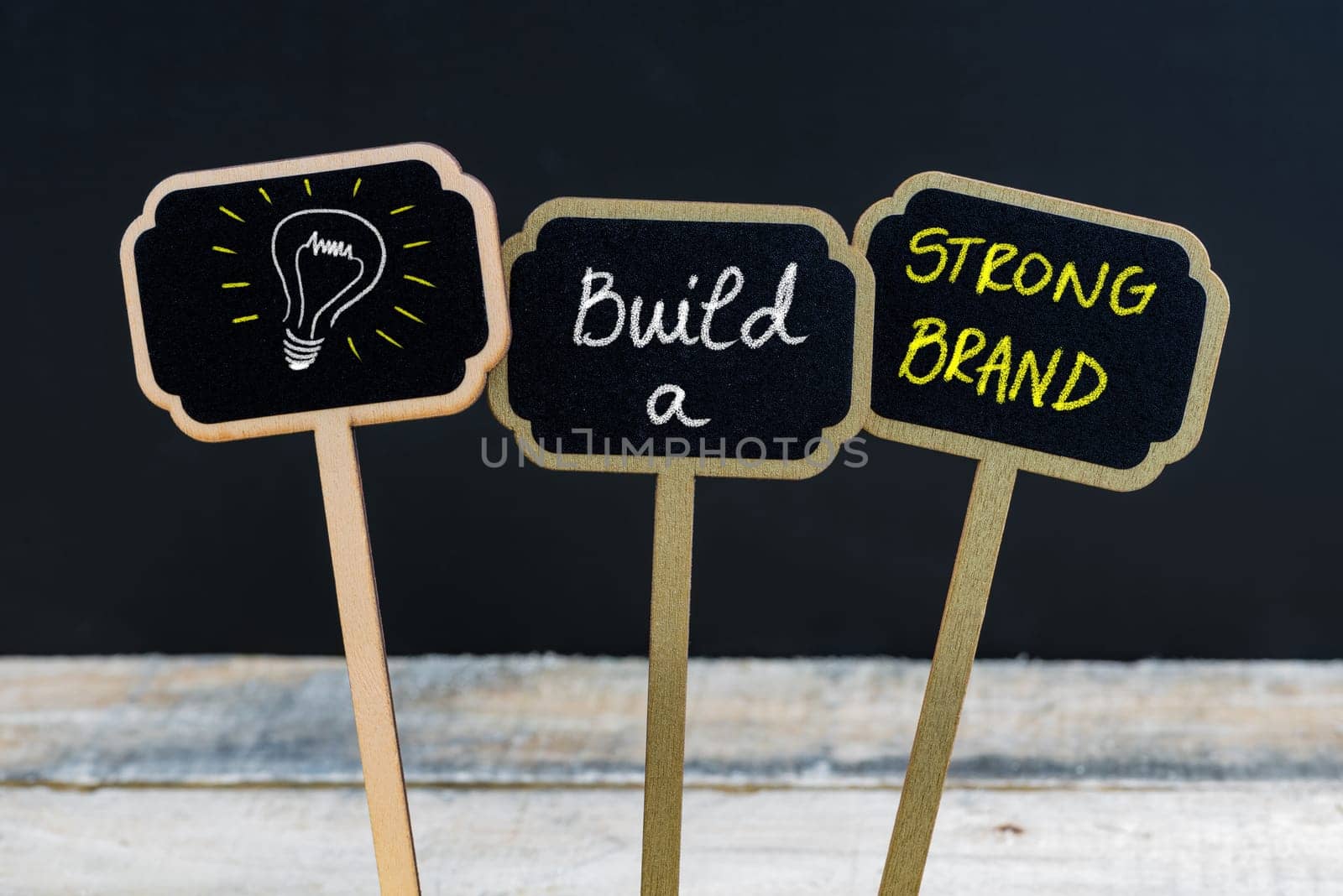 Concept message BUILD A STRONG BRAND and light bulb as symbol for idea written with chalk on wooden mini blackboard labels, defocused chalkboard and wood table in background