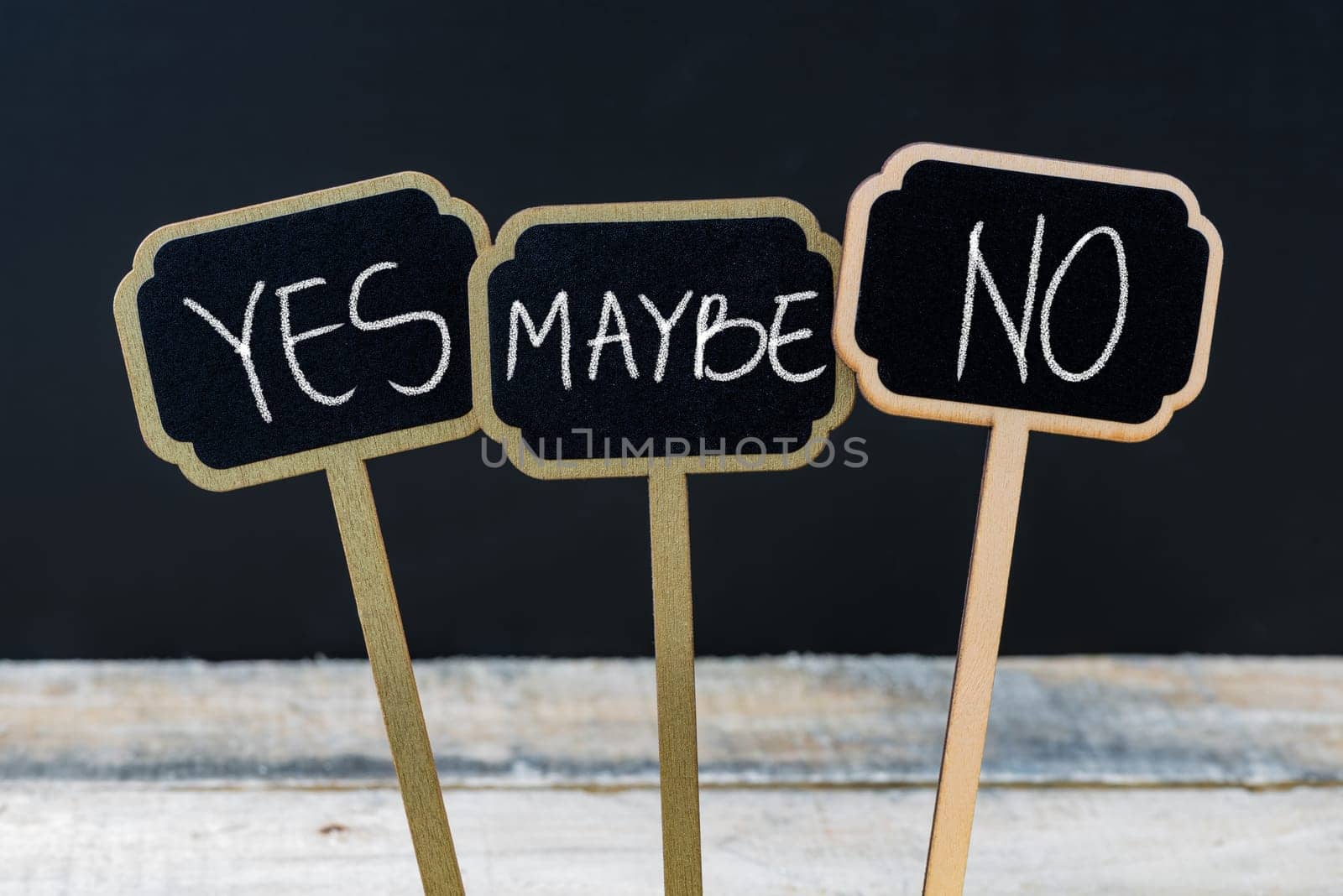 Concept message YES MAYBE NO written with chalk on wooden mini blackboard labels, defocused chalkboard and wood table in background