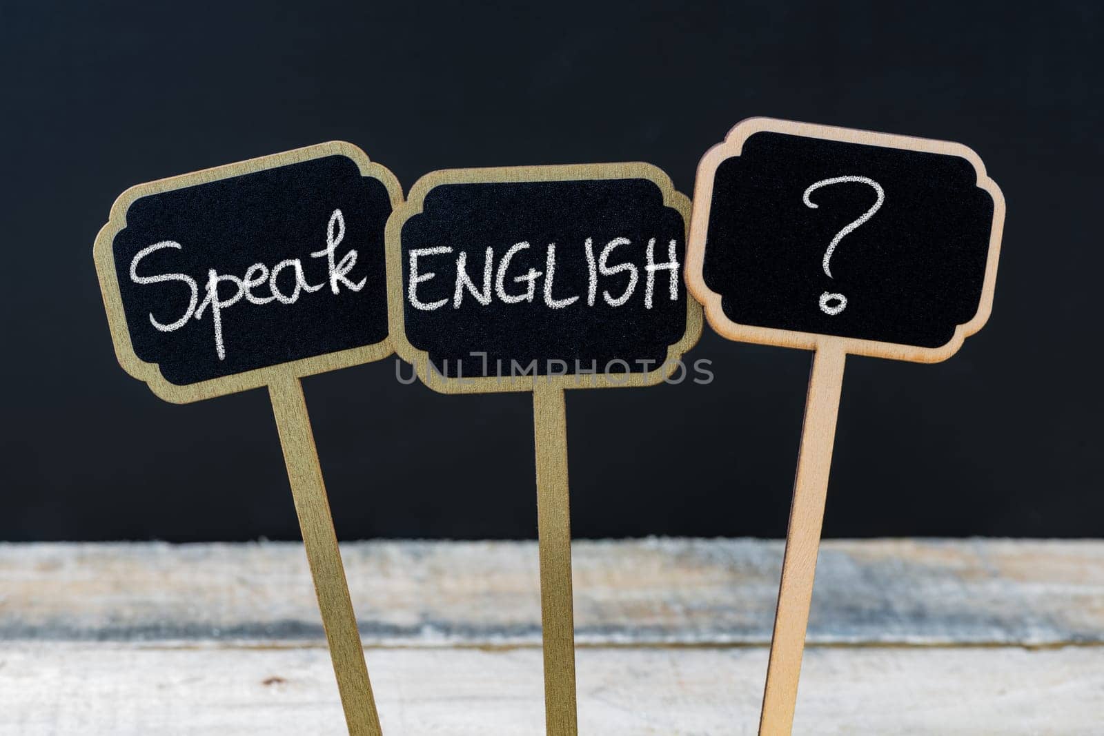Concept message SPEAK ENGLISH ? written with chalk on wooden mini blackboard labels, defocused chalkboard and wood table in background