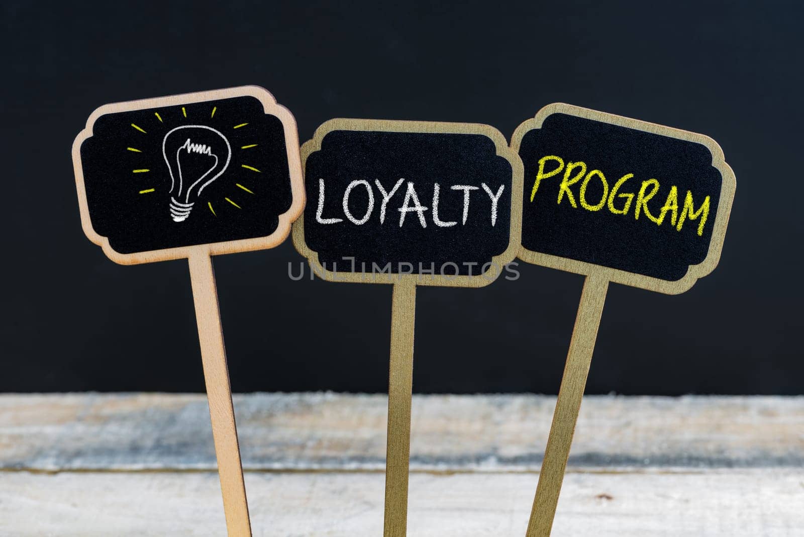 Concept message LOYALTY PROGRAM and light bulb as symbol for idea written with chalk on wooden mini blackboard labels, defocused chalkboard and wood table in background