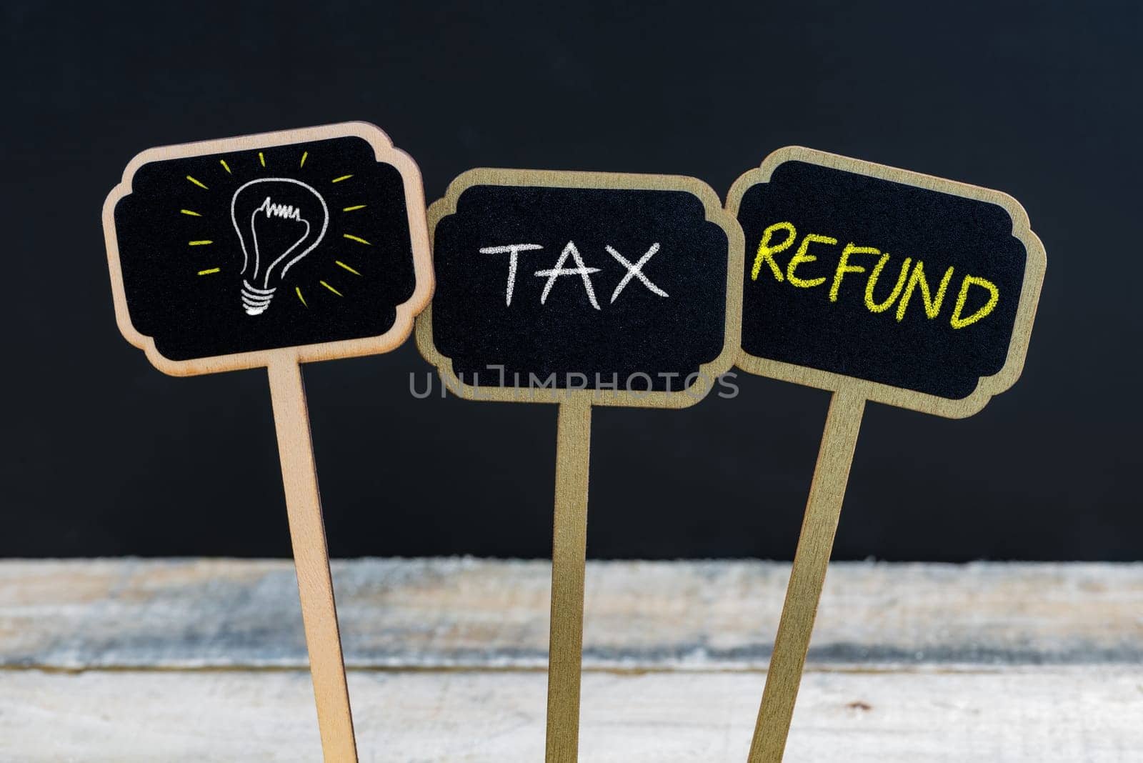 Concept message TAX REFUND and light bulb as symbol for idea written with chalk on wooden mini blackboard labels, defocused chalkboard and wood table in background