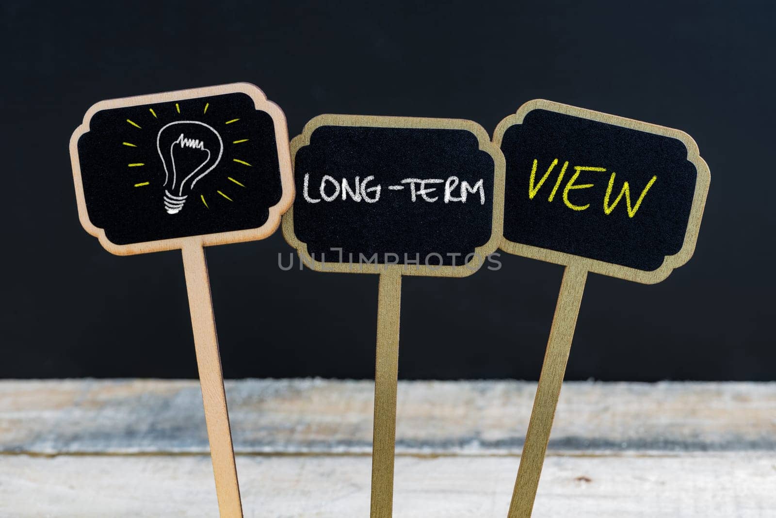Concept message LONG-TERM VIEW and light bulb as symbol for idea written with chalk on wooden mini blackboard labels, defocused chalkboard and wood table in background