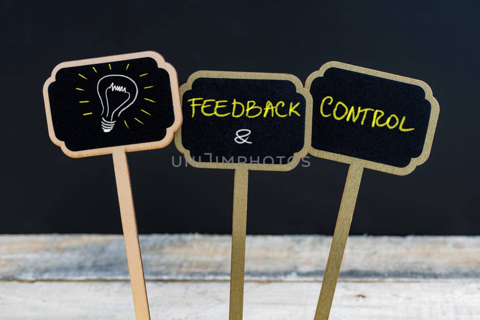 Concept message FEEDBACK and CONTROL and light bulb as symbol for idea written with chalk on wooden mini blackboard labels, defocused chalkboard and wood table in background