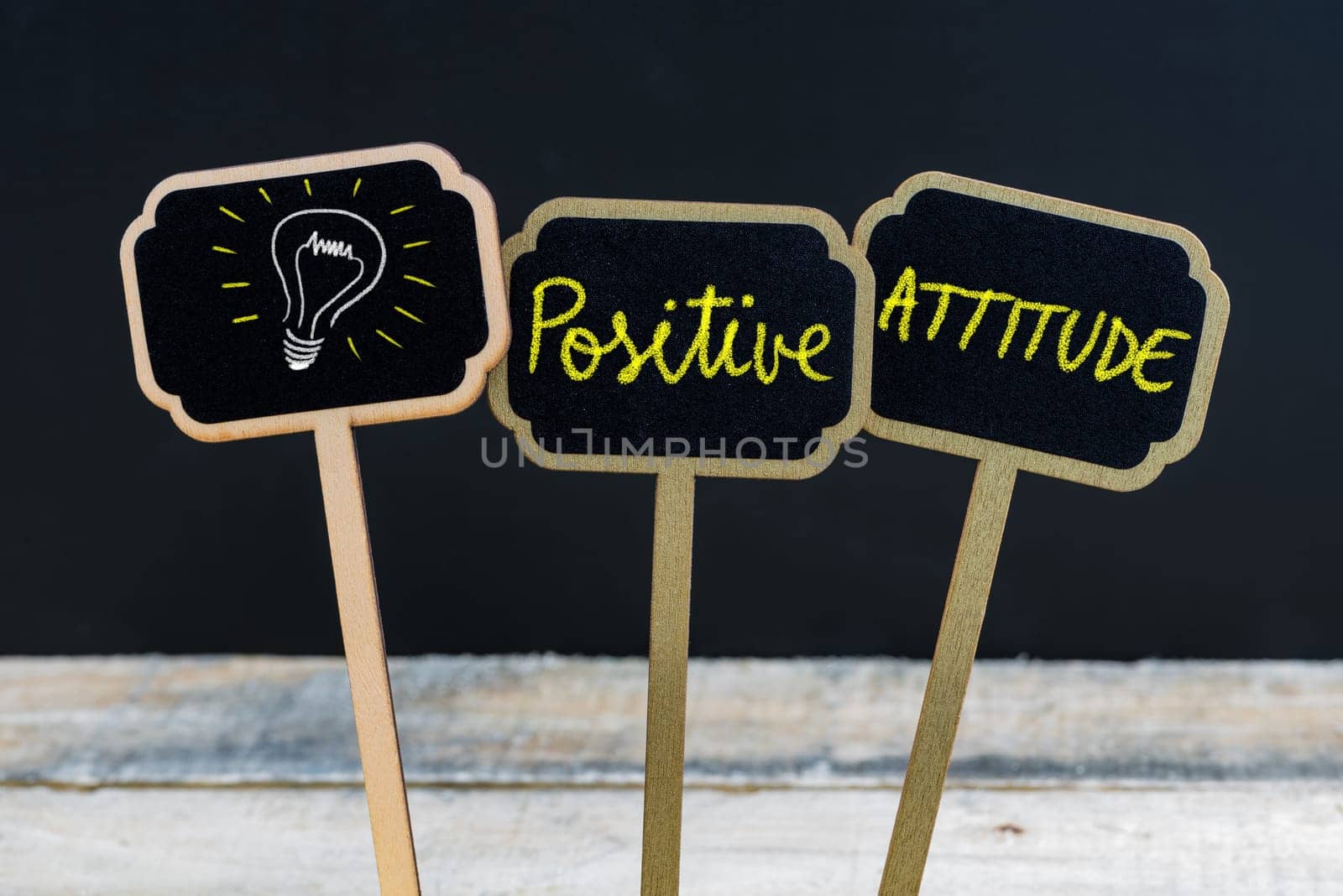 Concept message POSITIVE ATTITUDE and light bulb as symbol for idea written with chalk on wooden mini blackboard labels, defocused chalkboard and wood table in background