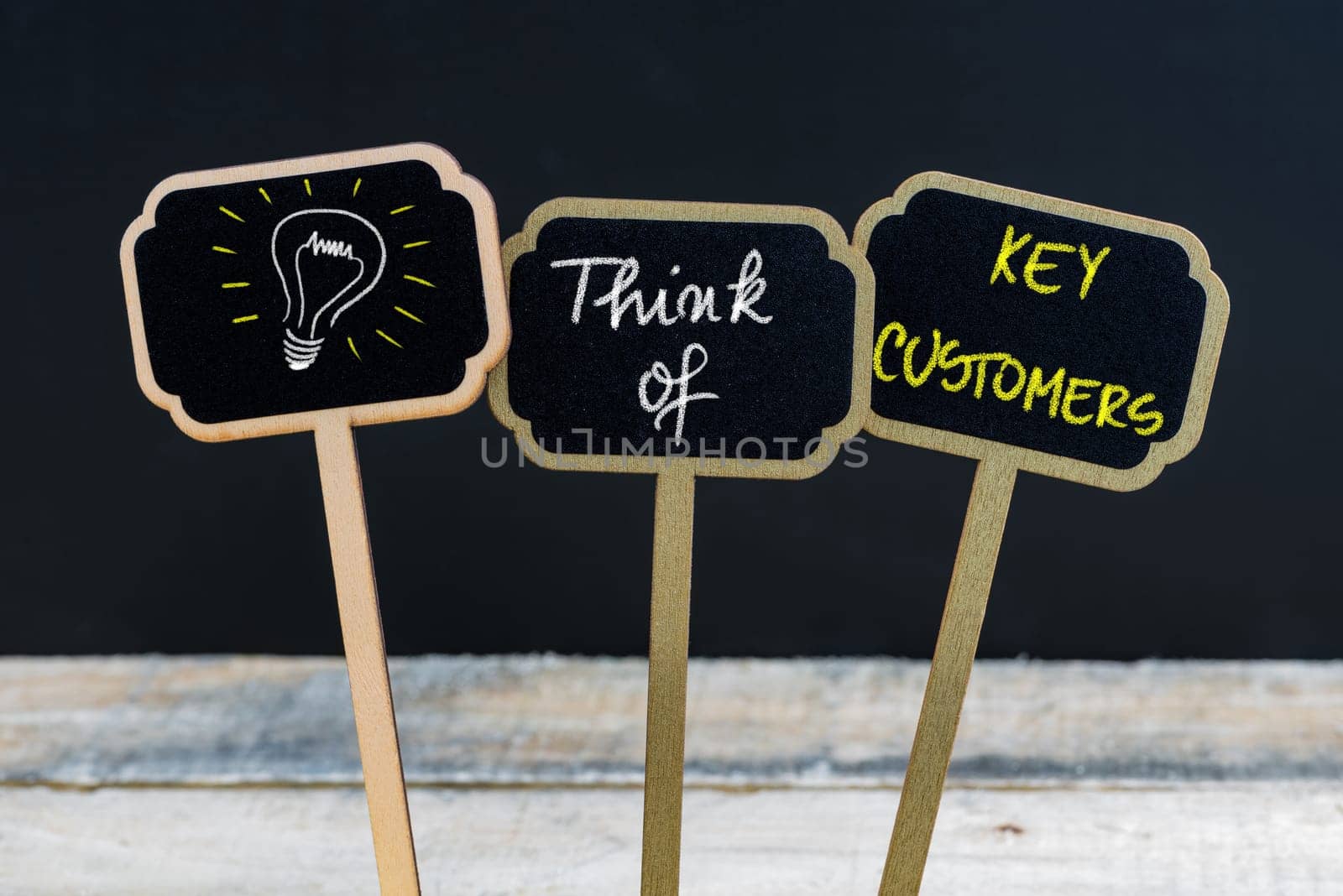 Concept message THINK OF KEY CUSTOMERS and light bulb as symbol for idea written with chalk on wooden mini blackboard labels, defocused chalkboard and wood table in background
