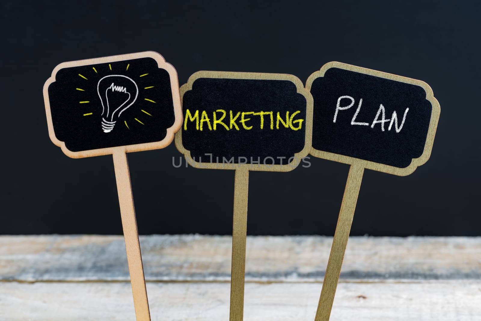 Concept message MARKETING PLAN and light bulb as symbol for idea written with chalk on wooden mini blackboard labels, defocused chalkboard and wood table in background