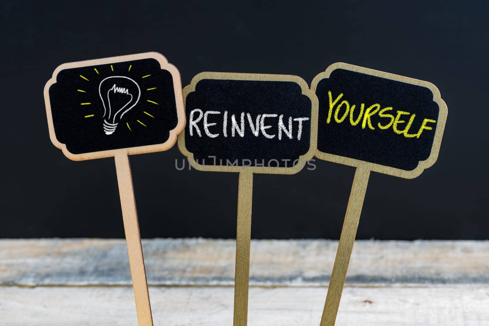 Concept message REINVENT YOURSELF and light bulb as symbol for idea written with chalk on wooden mini blackboard labels, defocused chalkboard and wood table in background