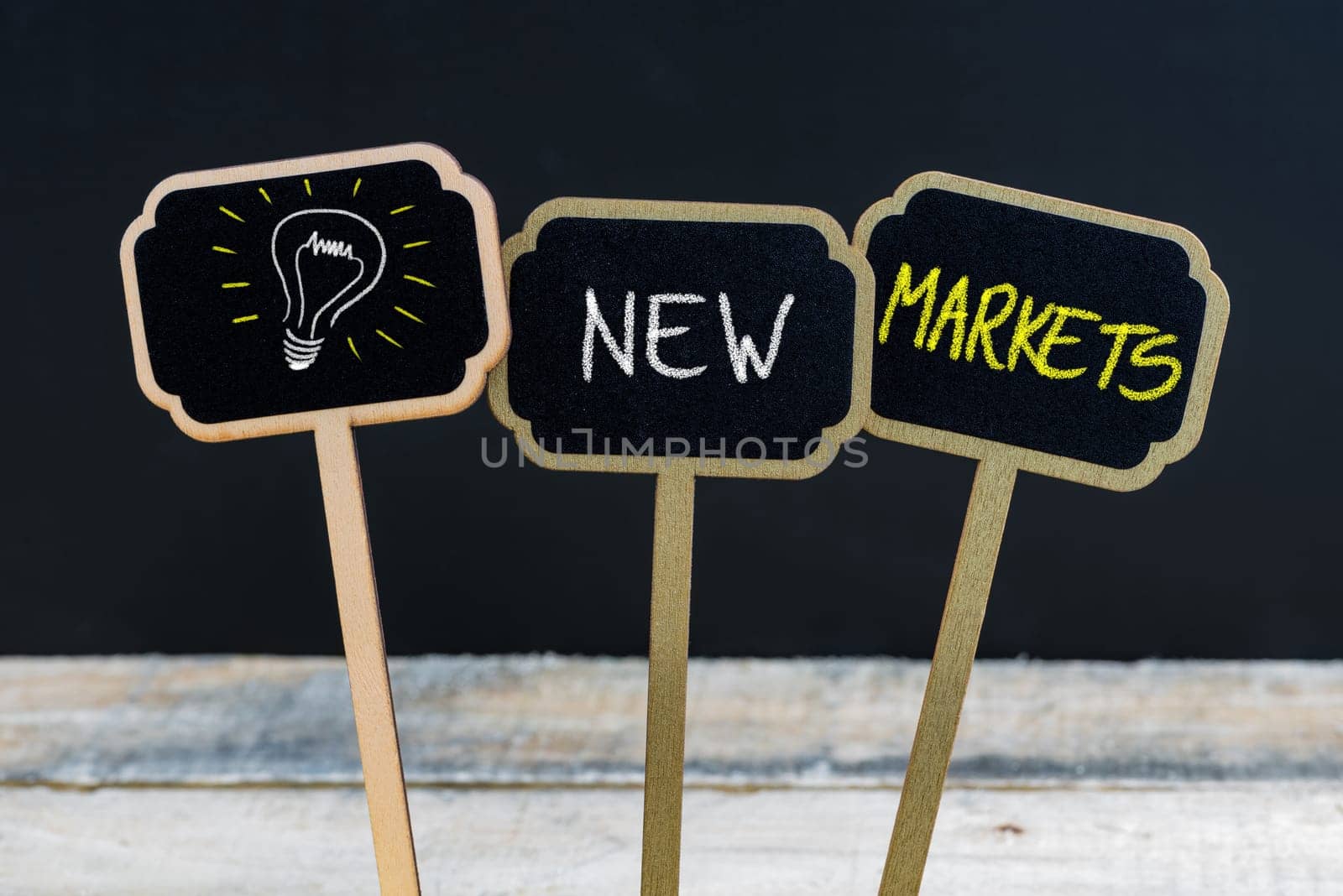 Concept message NEW MARKETS and light bulb as symbol for idea written with chalk on wooden mini blackboard labels, defocused chalkboard and wood table in background